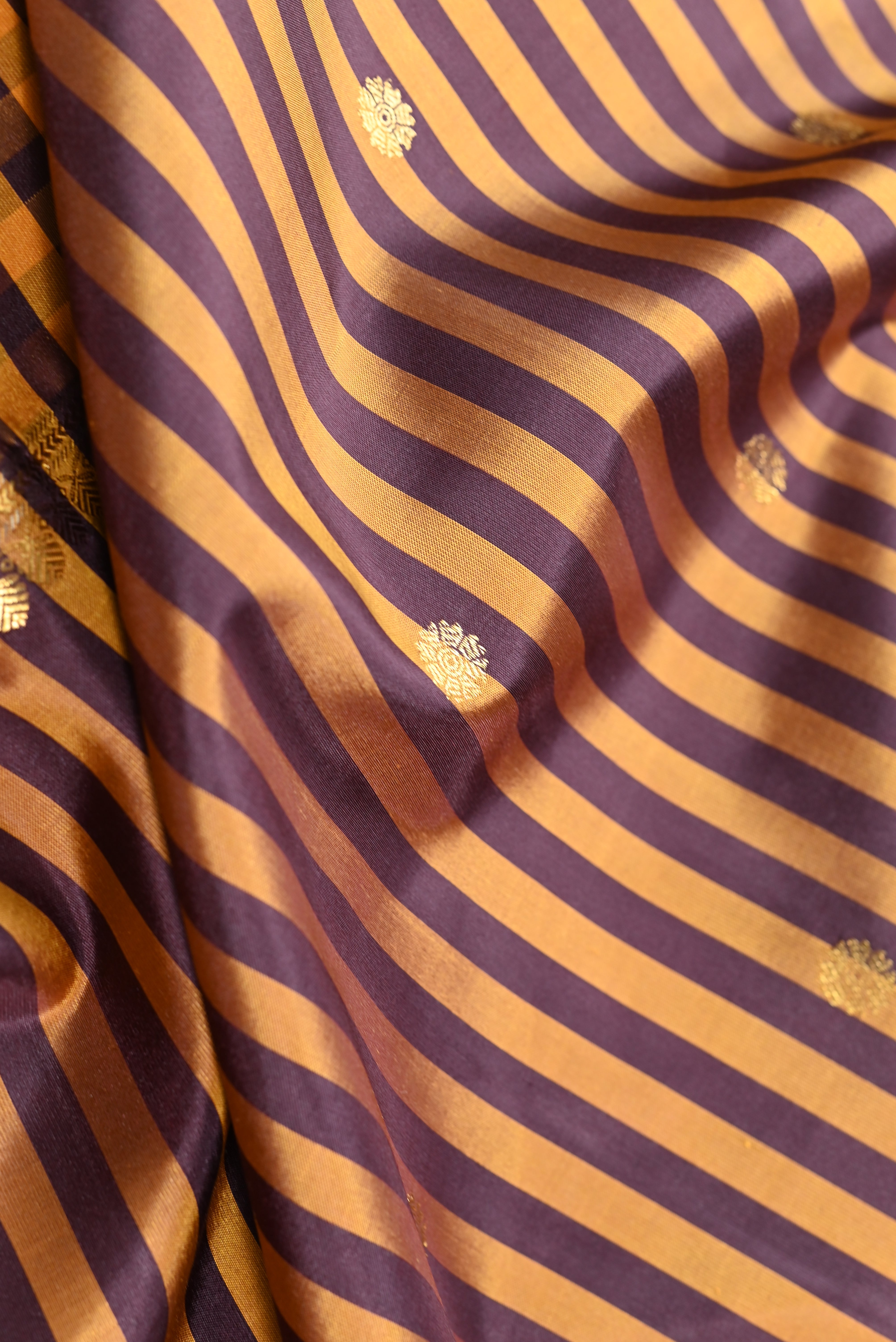 Close-up of a draped fabric with alternating wide and narrow diagonal stripes in warm mustard-gold and deep purple, accented with small metallic gold floral motifs.