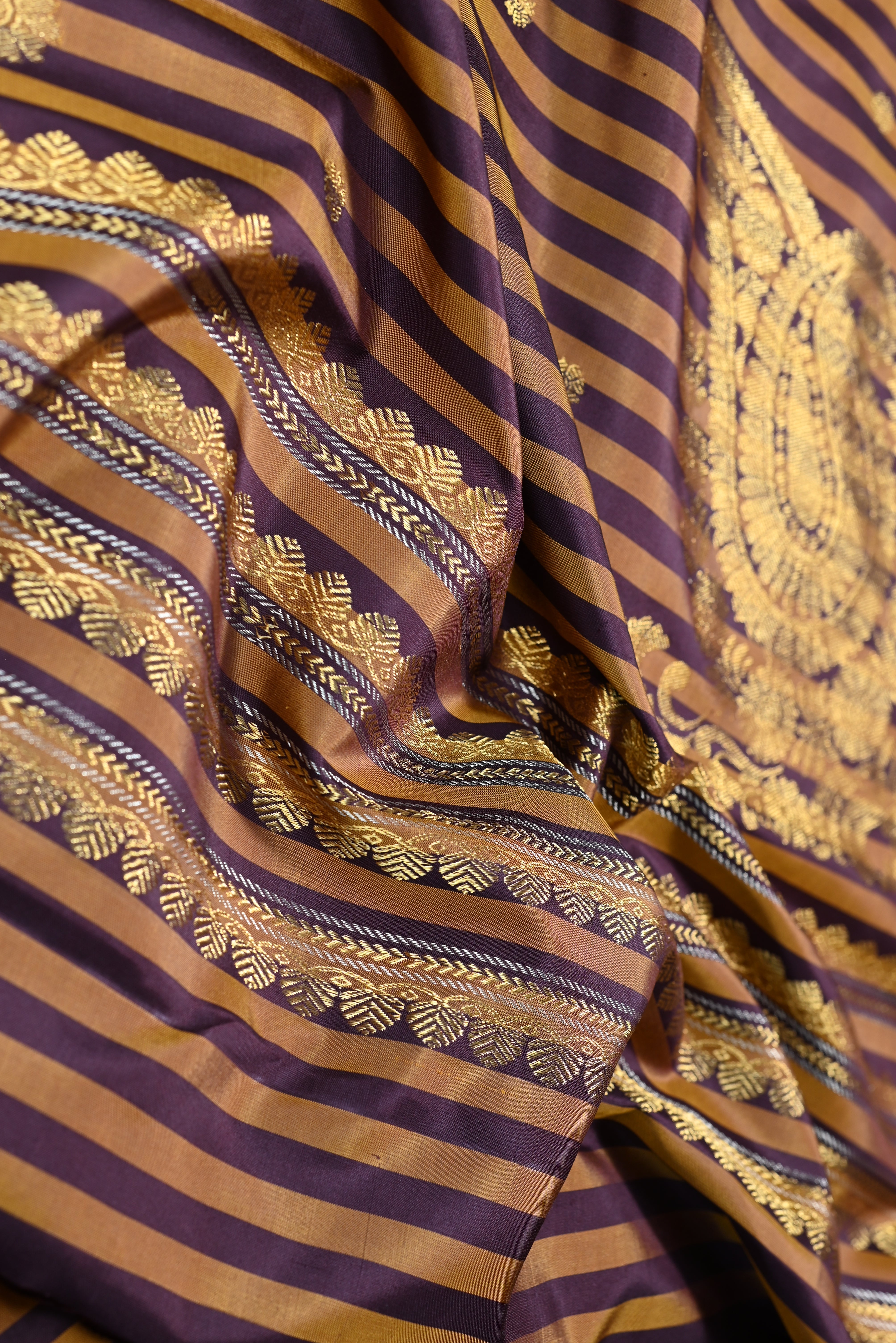 A close-up of a draped fabric with vertical stripes in deep purple and warm gold, decorated with intricate gold woven floral and geometric patterns.